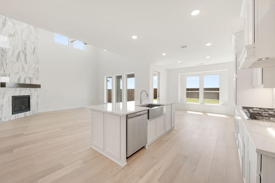 Open kitchen with white island, farmhouse sink, and light hardwood floors, open to living area with marble fireplace.