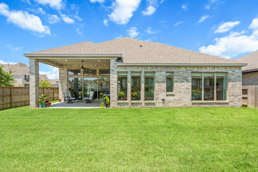 This photo showcases the backyard of a modern single-story home with a brick exterior. It features a spacious covered patio with outdoor seating, ideal for relaxation and entertaining. Complete with a well-maintained lawn. Large windows offer plenty of natural light inside the home.