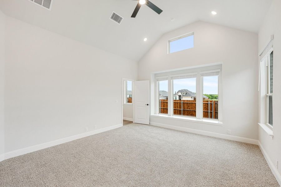 Empty bedroom with vaulted ceiling, ceiling fan, carpet flooring, and large windows overlooking backyard fence