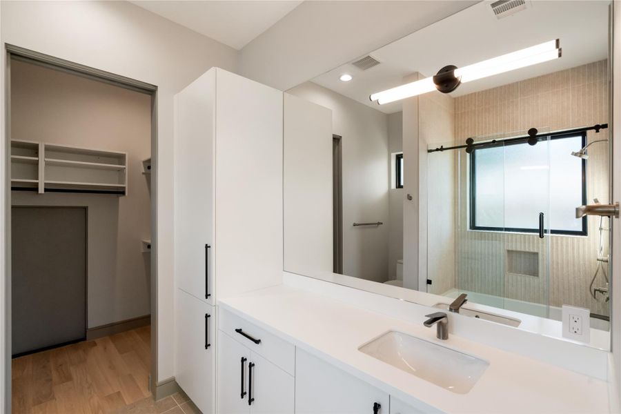 This modern bathroom features a sleek design with a spacious vanity, large mirror, and ample storage. It includes a glass-enclosed shower and a separate closet area with built-in shelving, all in a neutral color palette for a clean and contemporary feel.