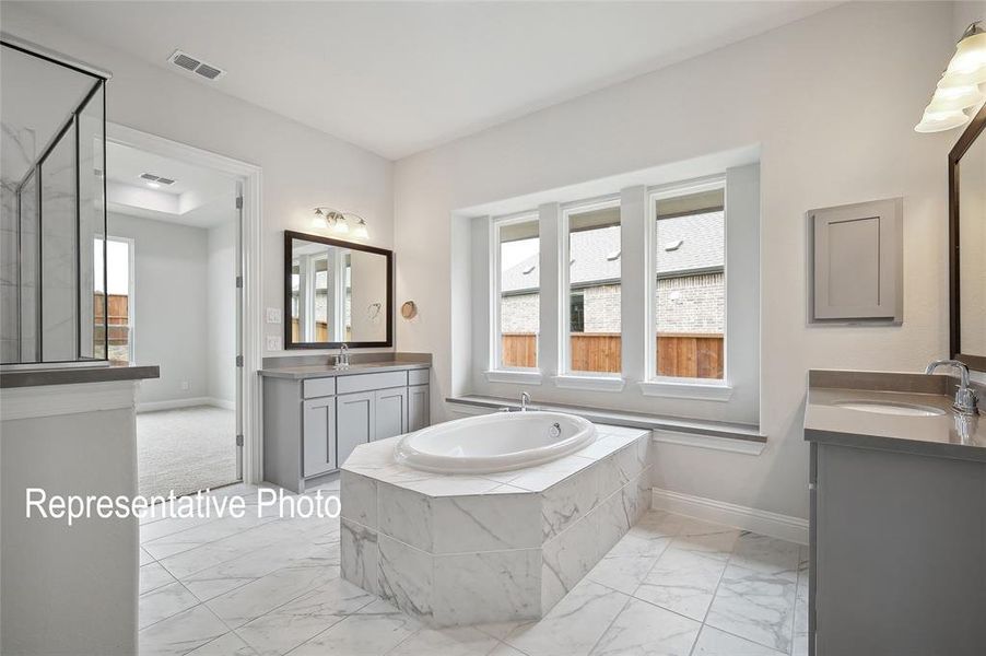 Spacious bathroom featuring a built-in soaking tub with marble-look tile surround, dual vanities with integrated sinks and quartz-look countertops, a separate glass-enclosed shower, and large windows above the tub