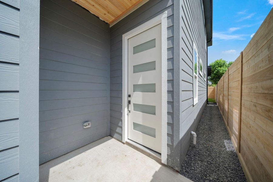 This photo shows a modern entrance with a sleek, white front door featuring frosted glass panels. The exterior is finished with horizontal gray siding, and there's a narrow, gravel side path leading to a fenced backyard. This photo shows a modern entrance with a sleek, white front door featuring frosted glass panels. The exterior is finished with horizontal gray siding, and there's a narrow, gravel side path leading to a fenced backyard.