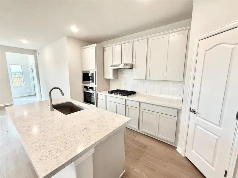 Kitchen with tasteful backsplash, light wood-style floors, a kitchen island with sink, light stone counters, and appliances with stainless steel finishes Kitchen with tasteful backsplash, light wood-style floors, a kitchen island with sink, light stone counters, and appliances with stainless steel finishes