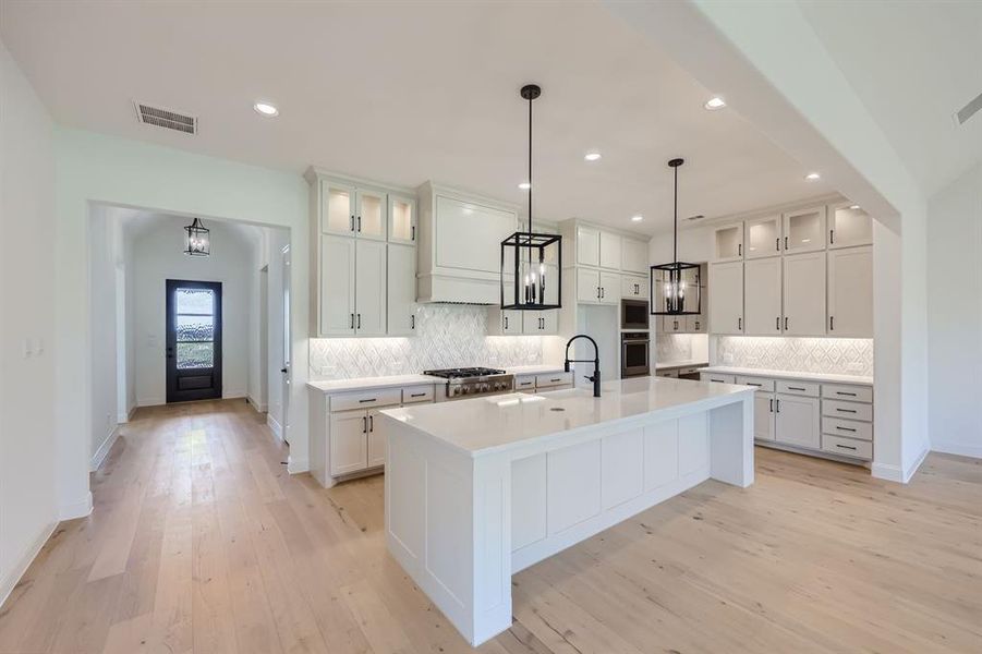 Kitchen featuring decorative light fixtures, decorative backsplash, appliances with stainless steel finishes, a center island with sink, and light hardwood / wood-style floors