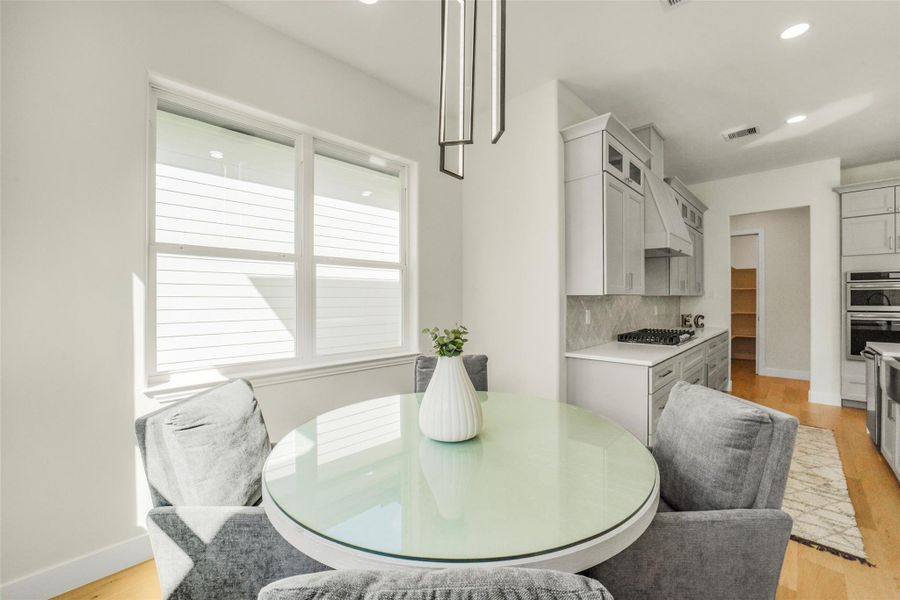 Bright and modern kitchen with light cabinetry and sleek appliances. Features a cozy breakfast nook with a glass-top table and plush chairs by a large window, providing ample natural light.