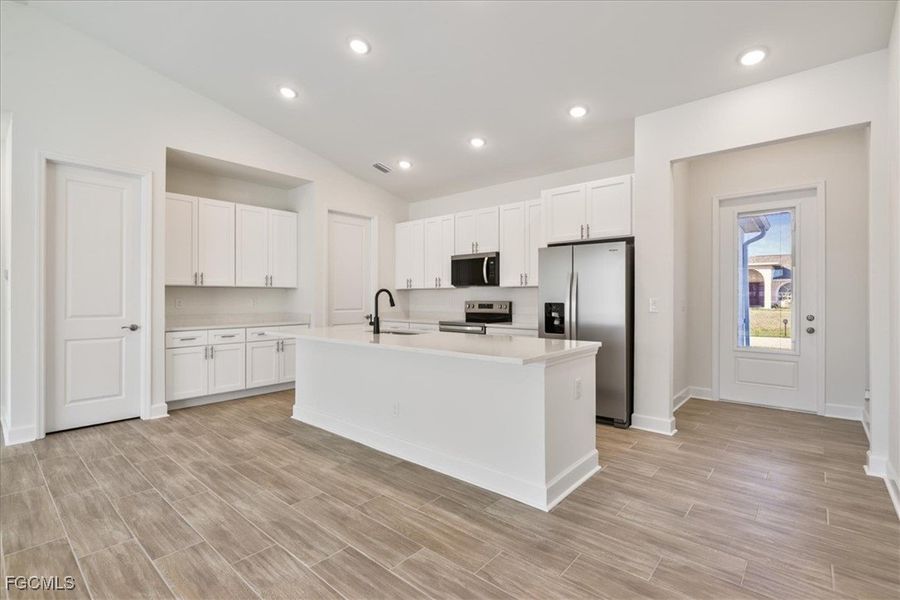 Kitchen featuring appliances with stainless steel finishes, vaulted ceiling, white cabinets, an island with sink, and wood tiled floors