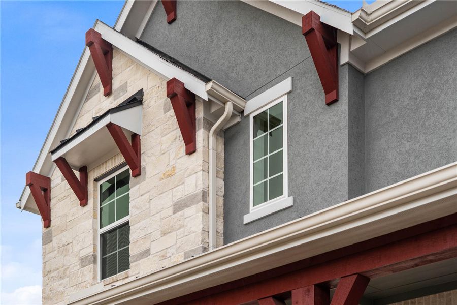 This striking modern elevation features a sophisticated blend of natural stone veneer and grey stucco, accented by bold, rust-colored timber corbels for a touch of rustic flair. Elegant gables and oversized windows with crisp white trim create an inviting, high-end look that ensures standout curb appeal for this new construction home.