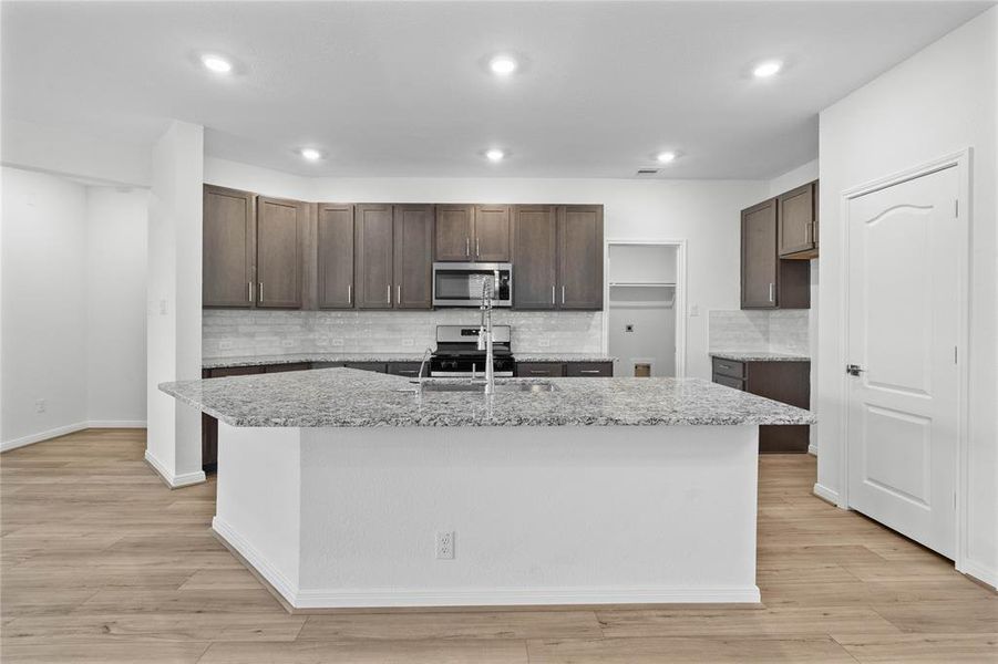 Another view of the kitchen featuring high ceilings, stained wood cabinets, granite countertops, SS appliances, modern tile backsplash, recessed lighting, large kitchen island with space for a breakfast bar, and a pantry all overlooking your huge family room.