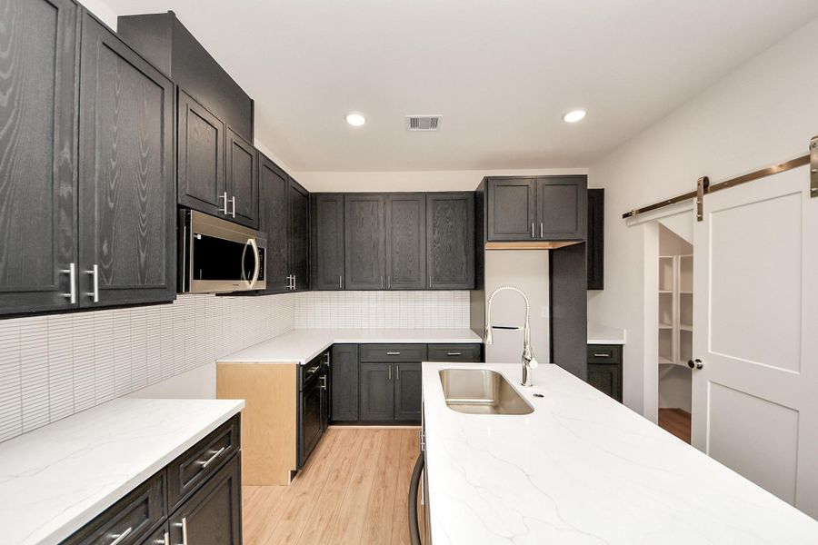 The V2 Floorplan – Kitchen. This sleek kitchen features dark cabinetry, quartz countertops, and a spacious island with a pull-down faucet. A sliding barn door opens to the walk-in pantry, while warm wood floors and stainless steel accents complete the modern design.