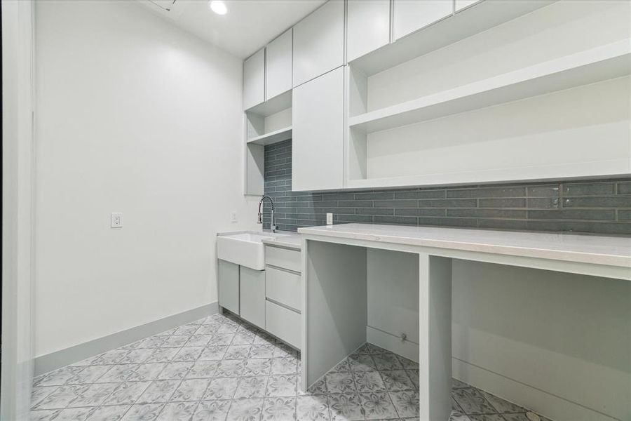 Featuring designer patterned ceramic tile flooring and Sherwin Williams Sea Salt-painted upper and lower cabinetry, this utility room offers a blue Zellige tile backsplash in a stacking pattern. Quartz countertops complement the space, which also includes washer and dryer connections and an apron front single basin sink.