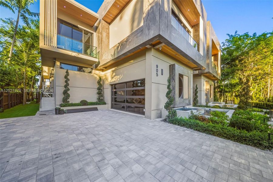 Wide paver motor court with glass-panel garage door, stone and stucco facade, spiral topiaries, recessed entry portico with wood-plank ceiling, circular porthole window, reflecting pool edge, and manicured landscape beds.