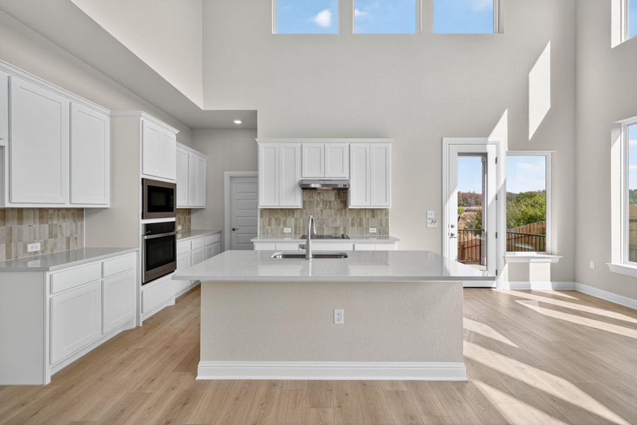 Kitchen with a towering ceiling, white cabinets, light stone countertops, light wood-type flooring, and an island with sink Kitchen with a towering ceiling, white cabinets, light stone countertops, light wood-type flooring, and an island with sink