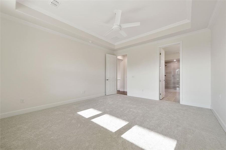 Unfurnished bedroom with ceiling fan, a raised ceiling, crown molding, ensuite bathroom, and light colored carpet