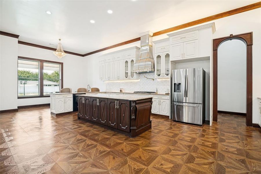 Expansive kitchen featuring a large central island with a stone countertop, white upper cabinetry, stainless steel appliances, and wood-finish flooring with a geometric pattern