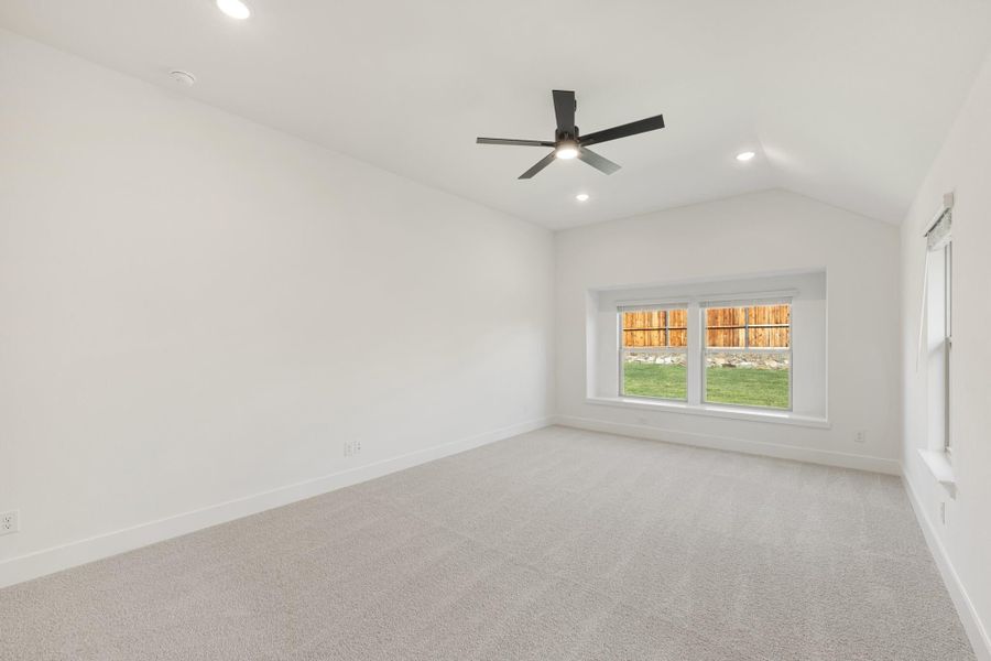 Empty bedroom with vaulted ceiling, black ceiling fan, carpet flooring, and a large window overlooking a backyard.