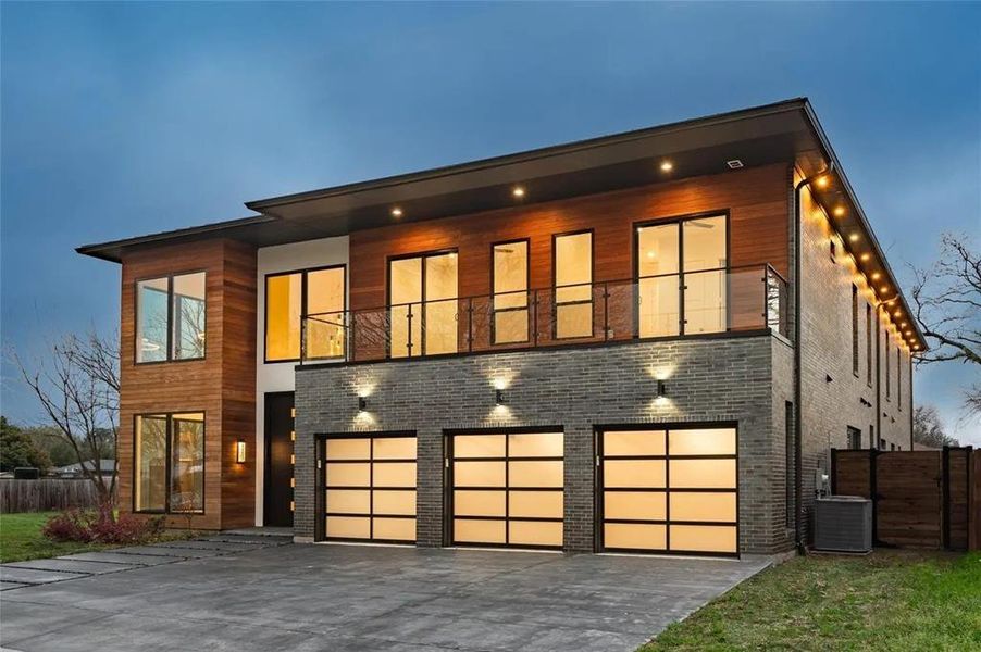 Modern exterior featuring wood paneling, dark brick accents, a three-car garage with frosted glass doors, a second-story balcony with glass railing, and a concrete drive Modern exterior featuring wood paneling, dark brick accents, a three-car garage with frosted glass doors, a second-story balcony with glass railing, and a concrete drive