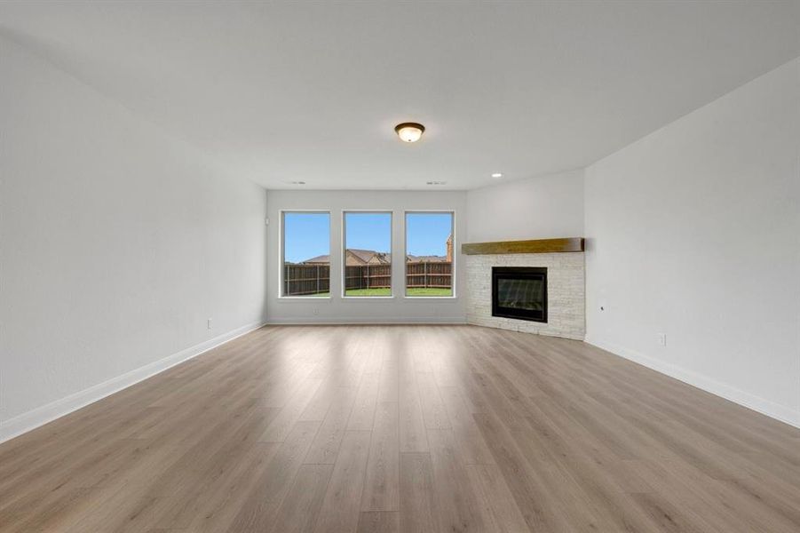 Spacious living area featuring light wood-finish flooring, white baseboards, and a contemporary fireplace with a light-colored stacked stone surround and a substantial wood mantel
