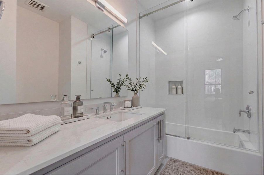 Modern full en-suite bath serving the first-floor bedroom, featuring Quartz countertops, undermount sink, sleek chrome fixtures, and a framed mirror. The glass-enclosed tub and shower combination includes a built-in niche and window for natural light, blending functionality with the elevated finishes found throughout this Nina Magon-designed residence. This photo has been virtually staged for visual reference.