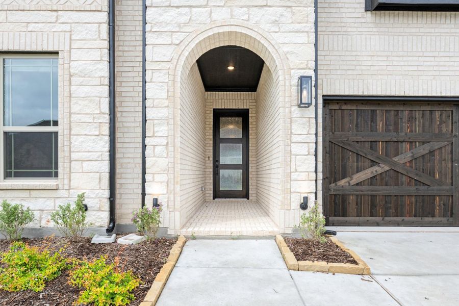 Home entrance with arched brick alcove, black front door, and rustic wood garage door beside landscaped walkway Home entrance with arched brick alcove, black front door, and rustic wood garage door beside landscaped walkway