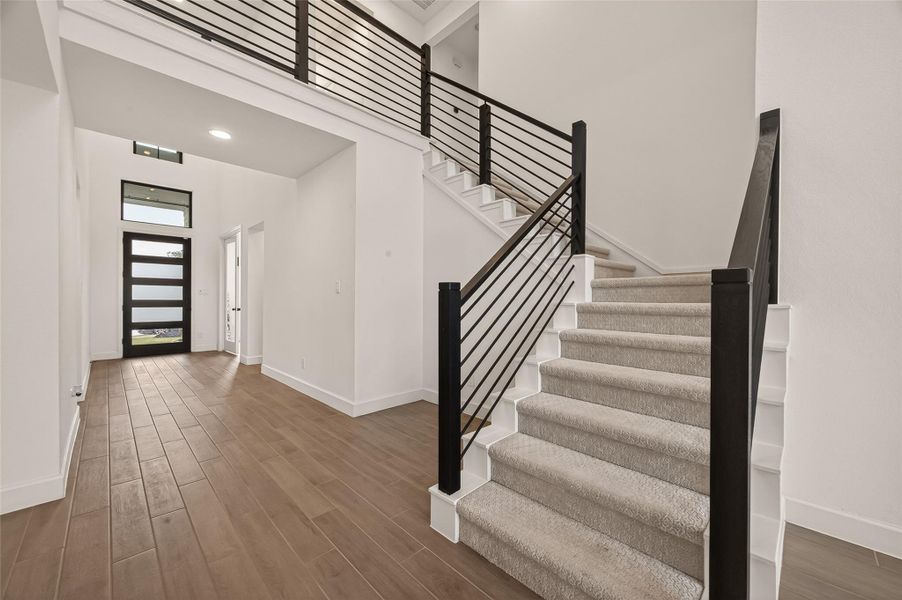 This photo showcases a modern entryway with a sleek staircase featuring dark railings. The space is bright with natural light from a large, contemporary front door and window. The flooring is a warm, wood-like tile, and the walls are painted white, creating a clean and inviting atmosphere.