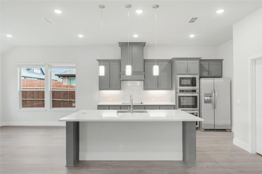 Kitchen with gray cabinets, white island, pendant lights, and stainless steel appliances on light wood floors