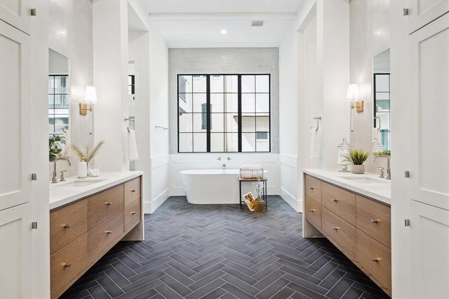 The magazine-worthy en suite owner's bath begins with herringbone Ann Sacks tile and floating waterfall double vanities in Forza Symphony Quartz and white oak. The bath features a Signature Hardware freestanding soaking tub with Kohler polished nickel plumbing hardware, set against a backdrop of grey plastered accent walls and aluminum windows, creating a luxurious and contemporary ambiance.