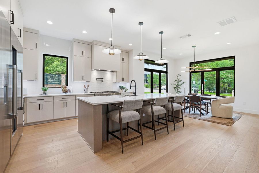 Open-concept kitchen featuring light wood-finish flooring, a central island with countertop seating, stainless steel appliances, light cabinetry, and large black-framed windows