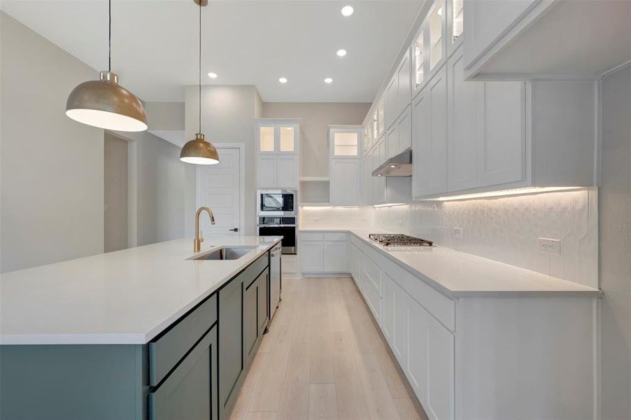 Kitchen with glass fronted cabinets, an island with sink, decorative light fixtures, light wood-type flooring, and stainless steel appliances Kitchen with glass fronted cabinets, an island with sink, decorative light fixtures, light wood-type flooring, and stainless steel appliances
