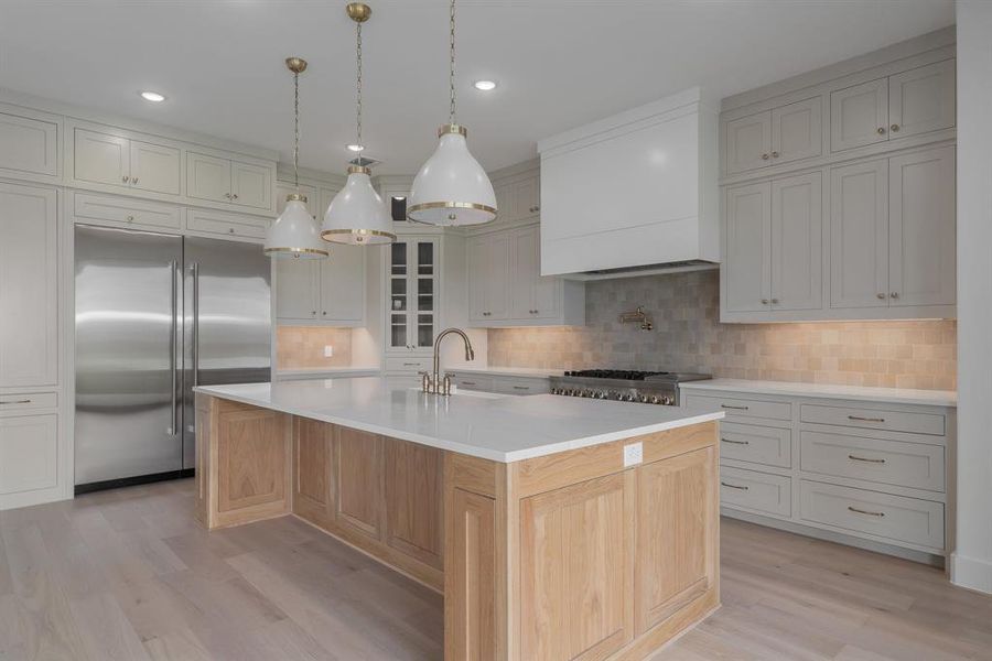 Kitchen featuring light hardwood / wood-style flooring, stainless steel built in fridge, an island with sink, custom exhaust hood, and decorative backsplash