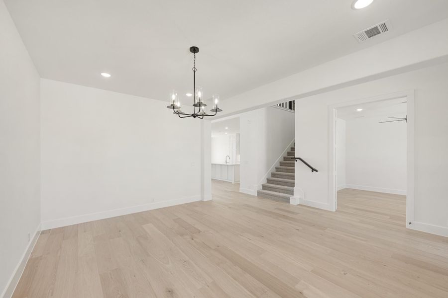 Open living area with light hardwood floors, black chandelier, white walls, and staircase visible in background.