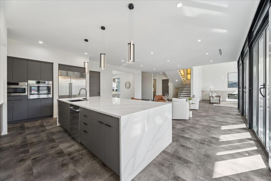 This modern home features an open-concept kitchen and living area with beautiful cabinetry, a large marble island, and pendant lighting. The space is bright, with floor-to-ceiling windows and a view of the staircase leading to the upper level.