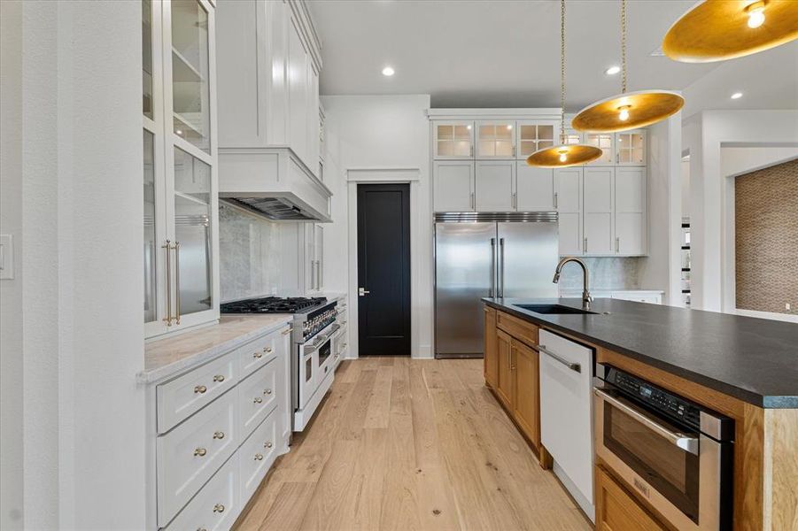Kitchen with high quality appliances, light wood-style flooring, a kitchen island with sink, hanging light fixtures, and glass insert cabinets