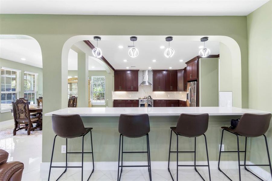 This photo showcases a modern kitchen with dark wood cabinets, stainless steel appliances, and a spacious countertop with bar seating for four. The archway design and pendant lighting add elegance, and the open layout connects to a bright dining area with ample natural light.