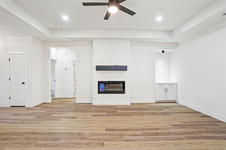 Unfurnished living room with a glass covered fireplace, light wood-style floors, a tray ceiling, a ceiling fan, and recessed lighting Unfurnished living room with a glass covered fireplace, light wood-style floors, a tray ceiling, a ceiling fan, and recessed lighting
