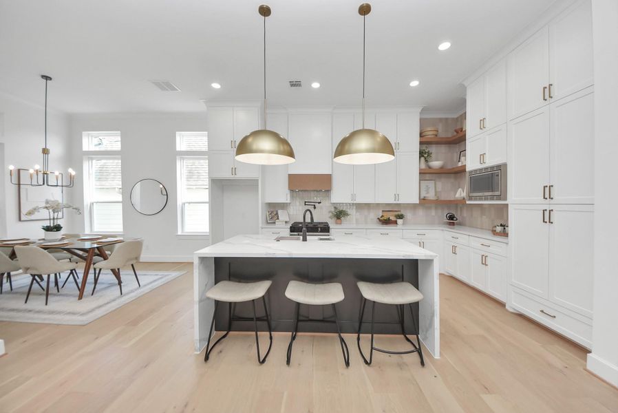 This modern kitchen features elegant white cabinetry, a large marble island with seating, and stylish pendant lighting. The open layout flows into a dining area with ample natural light, creating a bright and inviting space.