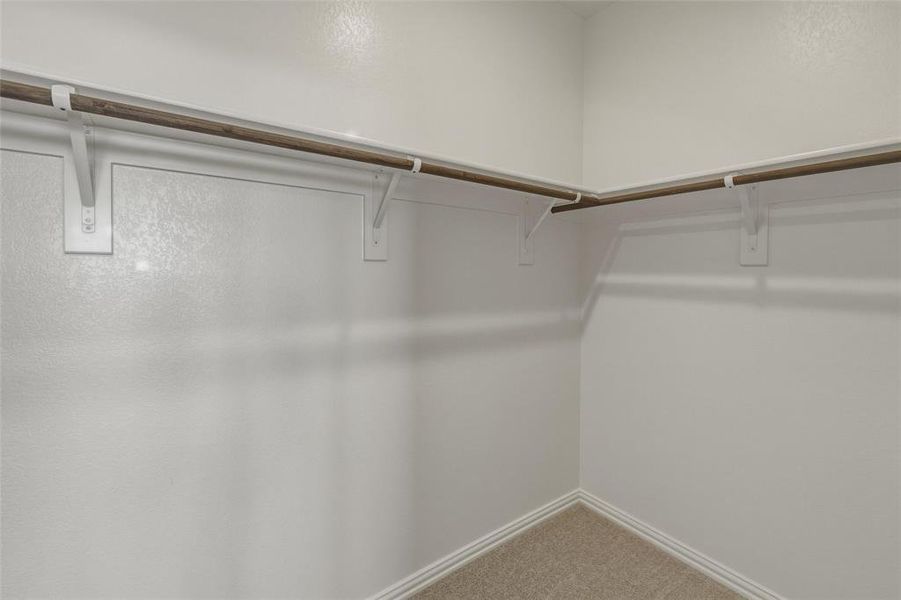 Walk-in closet featuring white walls, wood-finish hanging rods, white support brackets, and light-colored carpet Walk-in closet featuring white walls, wood-finish hanging rods, white support brackets, and light-colored carpet