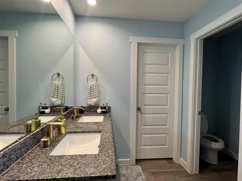 This bathroom features a modern design with a double-sink vanity and granite countertop. It includes a spacious glass-enclosed shower with white subway tiles, a separate toilet area, and wood-look flooring. The soft blue walls and recessed lighting create a bright, inviting atmosphere.