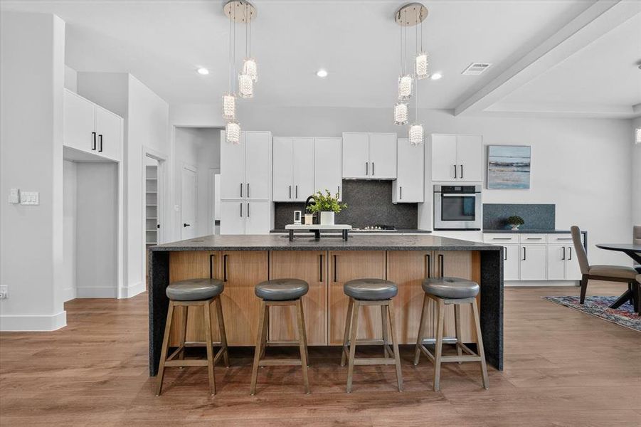 Spacious kitchen featuring a large island with seating, white shaker-style cabinetry, dark tile backsplash, stainless steel built-in appliances, and wood-finish flooring