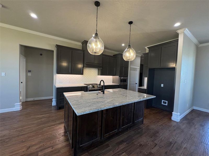 Kitchen featuring light stone countertops, pendant lighting, ornamental molding, a center island with sink, and dark wood-style floors