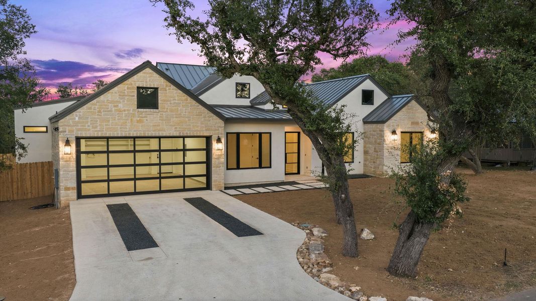 Mature oak trees frame a modern stone facade beneath a standing-seam metal roof.