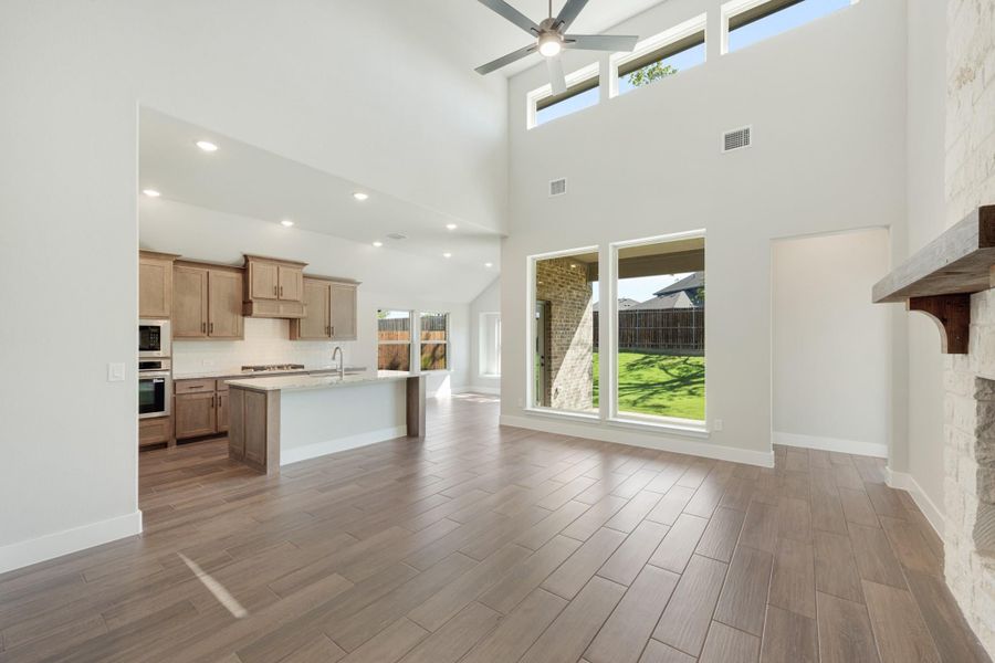 Open-concept living area with high ceilings, wood-look floors, and kitchen with island and stone fireplace mantel visible Open-concept living area with high ceilings, wood-look floors, and kitchen with island and stone fireplace mantel visible