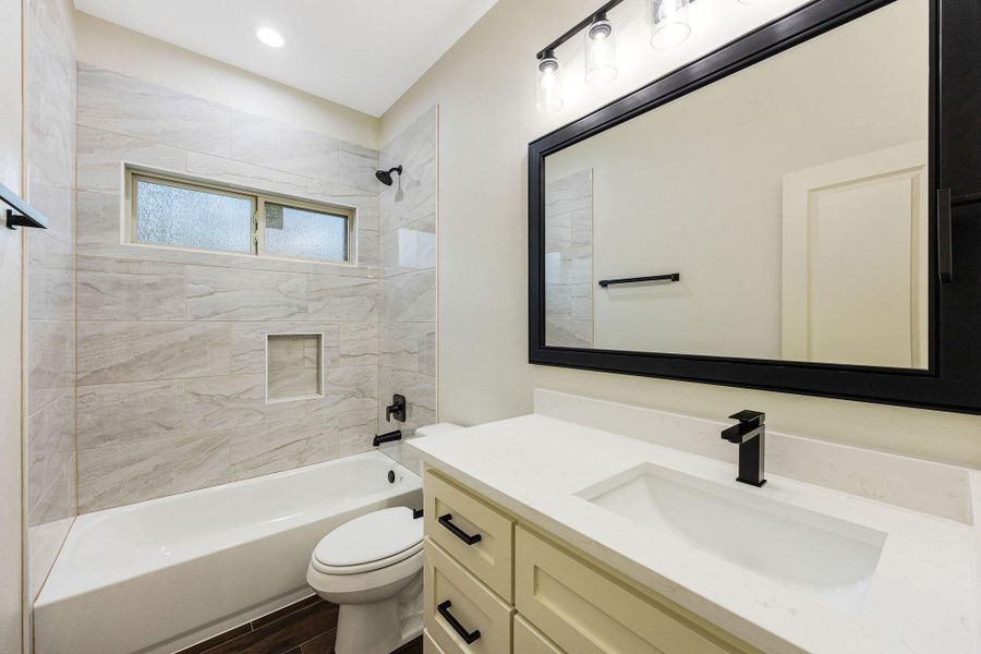 Modern secondary bathroom featuring a sleek vanity with a black-framed mirror, white countertop, and black fixtures. The shower-tub combo has elegant tile work with a large window for natural light.