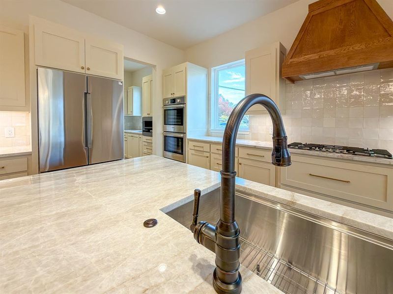 Spacious kitchen featuring a large island with a stainless steel farmhouse sink and bronze-finish faucet, light-toned cabinetry, stainless steel appliances including a built-in refrigerator and double oven, and a wood-paneled range hood over a gas cooktop