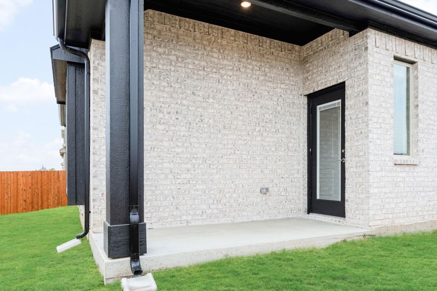 Covered back patio with white brick exterior, black door, concrete slab, and wood privacy fence