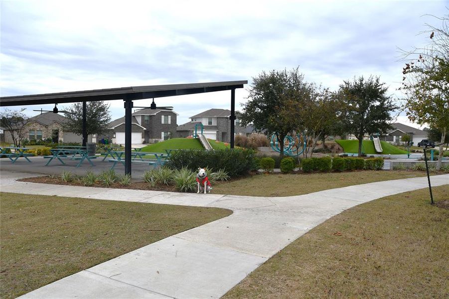 This photo shows a neighborhood park with picnic tables under a covered area, a playground with slides, and landscaped greenery. It’s a family-friendly environment with nearby houses, ideal for community gatherings and outdoor activities.