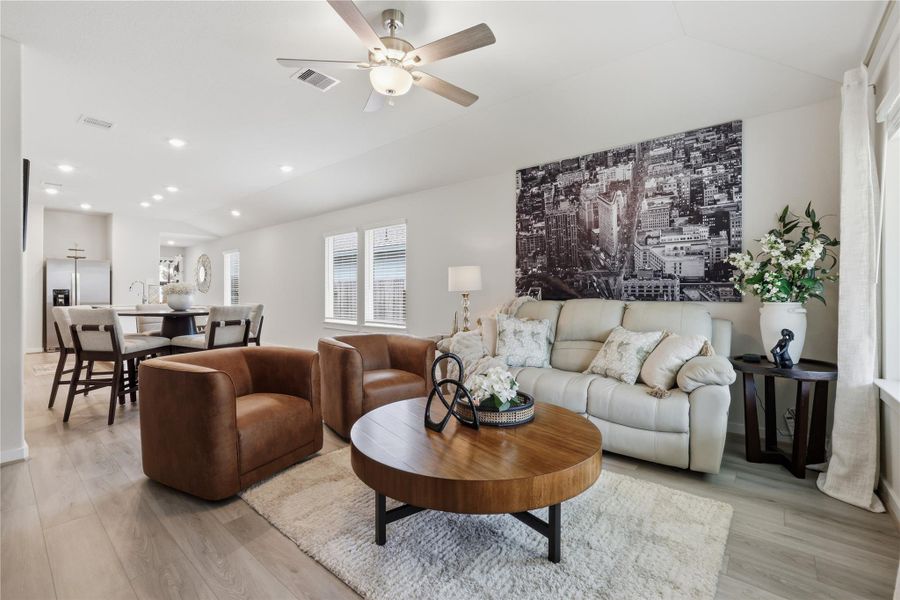 This modern living area features an open layout with stylish furnishings, including leather chairs and a cozy sofa. The space is accented by a large cityscape artwork and ample natural light from multiple windows. The room flows seamlessly into the dining and kitchen areas, creating a welcoming atmosphere.