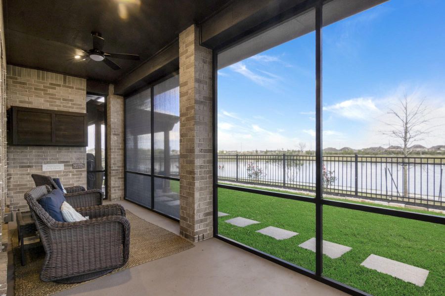 This photo showcases a cozy screened patio with a ceiling fan and outdoor TV, overlooking a serene lake. The area features a well-maintained lawn with stepping stones and a view through black metal fencing. Perfect for relaxing or entertaining with a scenic backdrop.