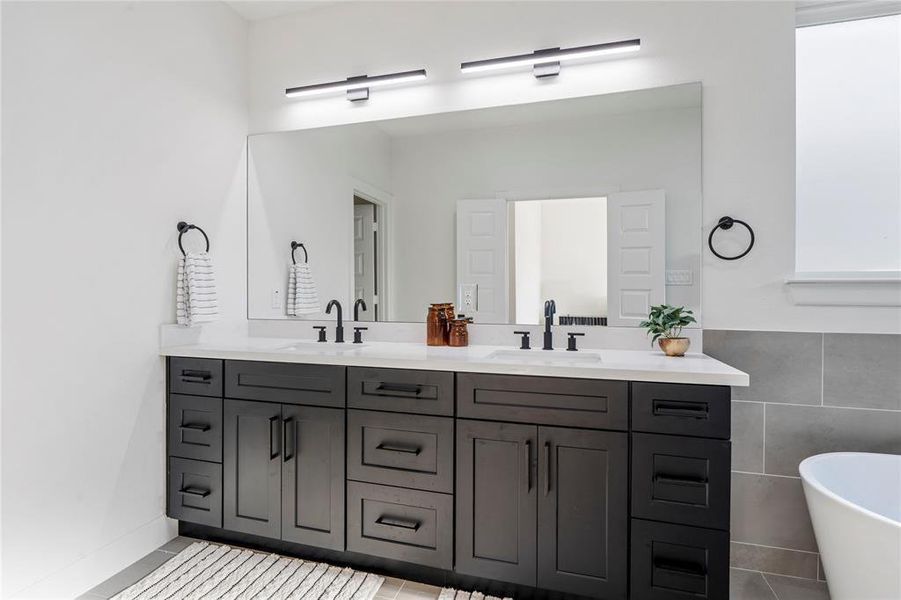 Sleek modern bathroom featuring a double vanity with dark cabinetry, elegant fixtures, and ample lighting, complemented by a large mirror and a freestanding bathtub. Sleek modern bathroom featuring a double vanity with dark cabinetry, elegant fixtures, and ample lighting, complemented by a large mirror and a freestanding bathtub.