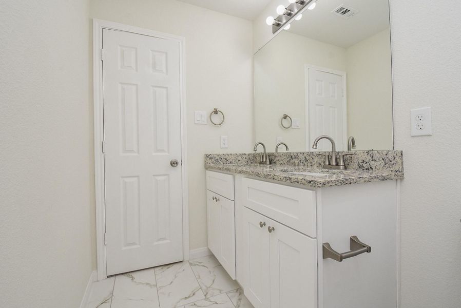 A modern bathroom with a white vanity, granite countertop, two sinks, a large mirror, and bright lighting above the mirror. A modern bathroom with a white vanity, granite countertop, two sinks, a large mirror, and bright lighting above the mirror.