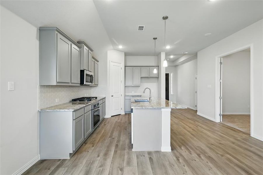 Kitchen with gray cabinetry, light stone countertops, decorative backsplash, and a center island with sink